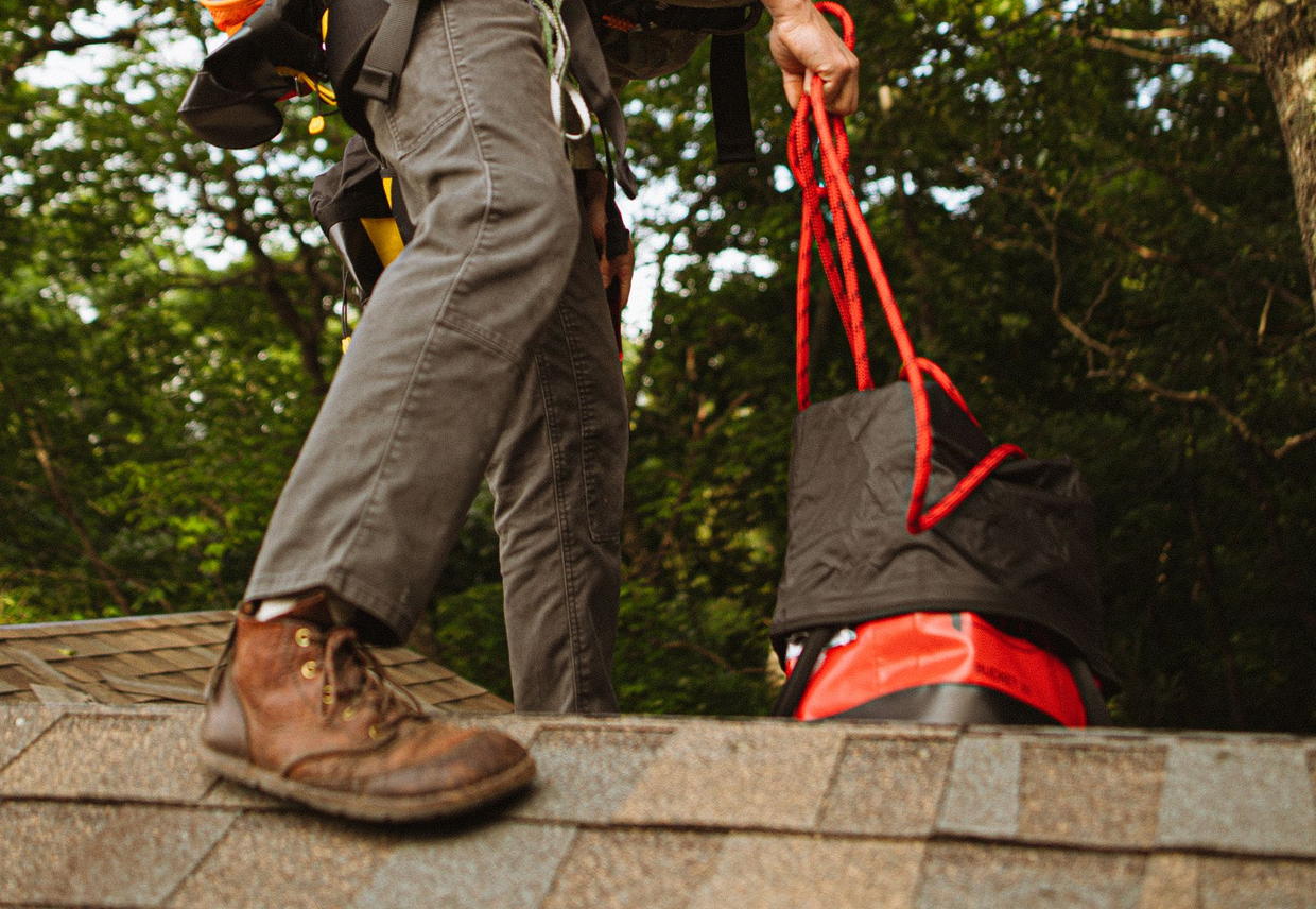 Licensed stock photograph: a roofer in worn leather boots and a fall harness, stepping across a shingled slope with a rope bag in hand.